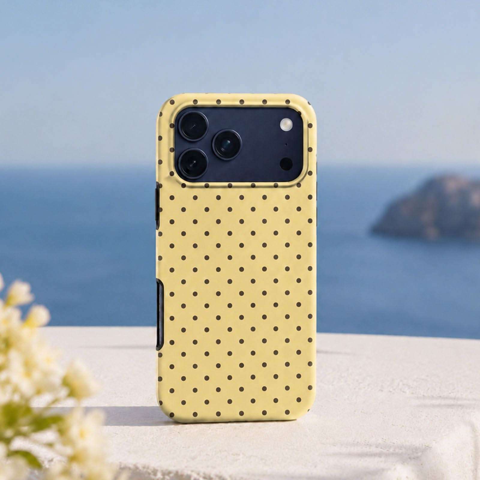 Yellow phone case with black polka dots, triple camera cutout, displayed on white surface with ocean background.