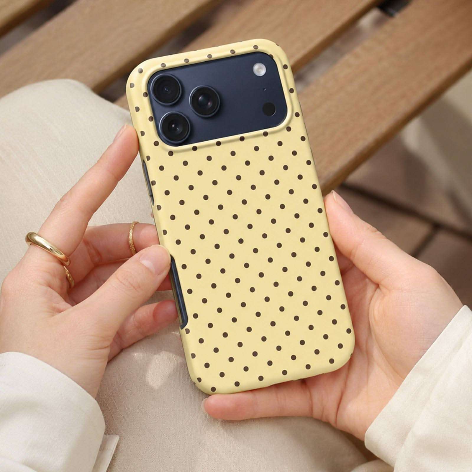 Pale yellow iPhone case with black polka dots and protective camera ring, held in hand.