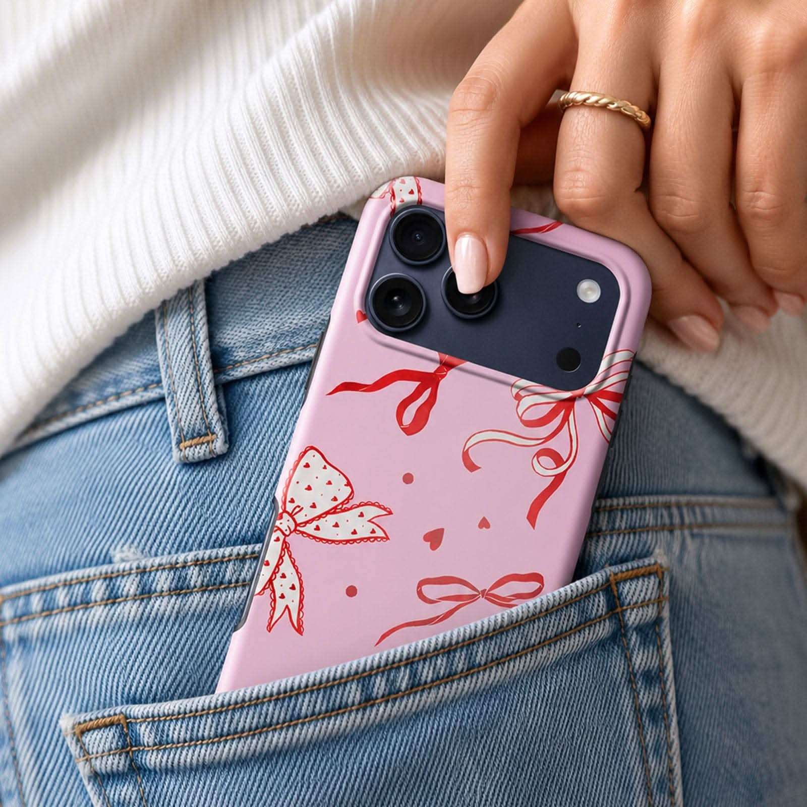 Pink phone case with red ribbon and bow design, displayed in jeans pocket with gold ring visible.